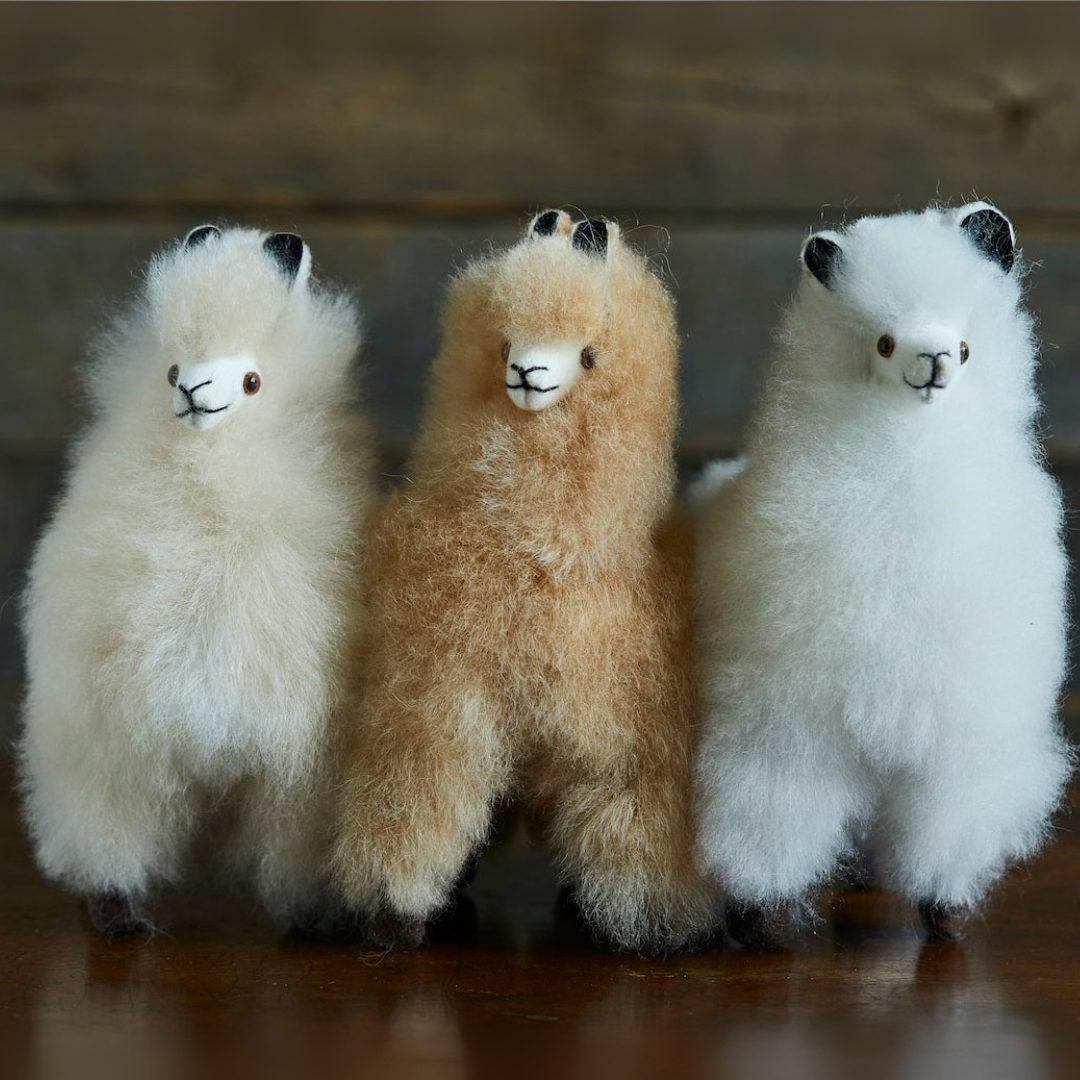 Three fluffy toy llamas in white, beige, and white on a wooden surface.